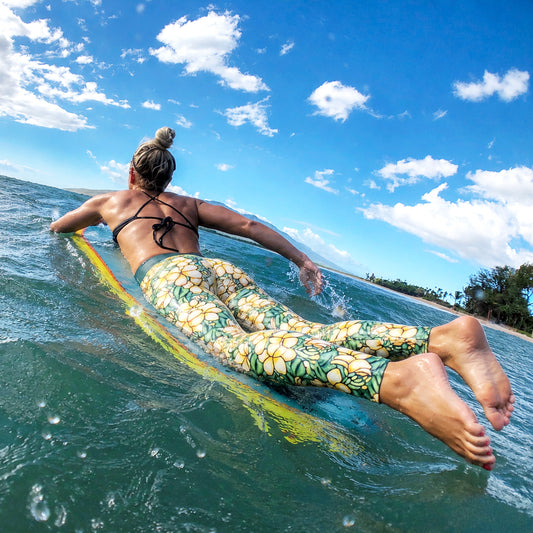 Person lying on a surfboard in the ocean with a clear blue sky wearing leggings with plumeria design.